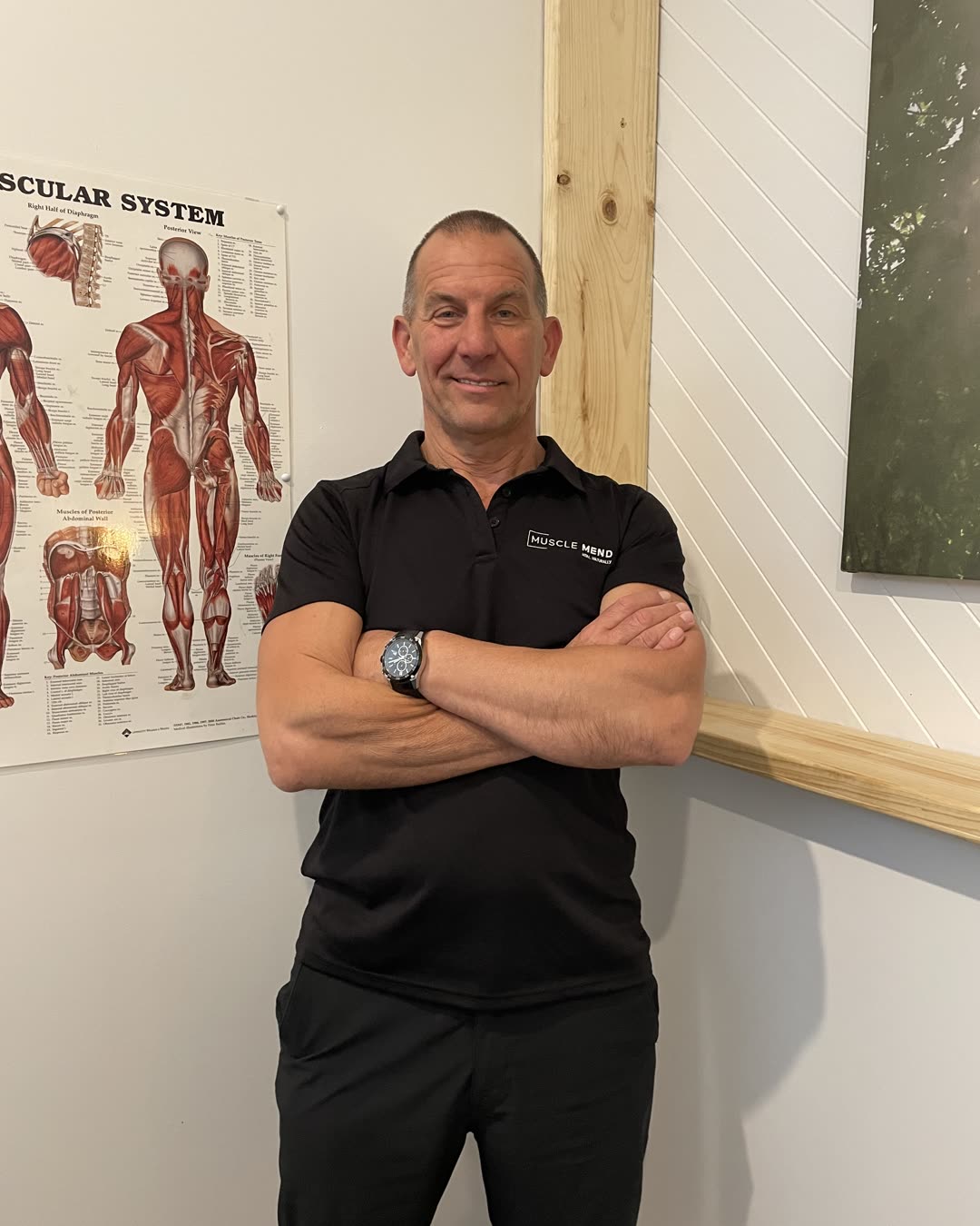 Mario, remedial massage therapist, smiling in the Muscle Mend treatment room in front of an anatomy chart